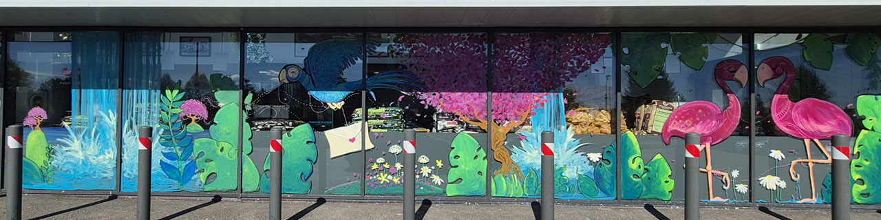 Photographie panoramique de la façade vitrée d'un supermarché ornée d'une grande fresque murale colorée. La scène de gauche montre des chutes d'eau et des feuillages tropicaux, le centre un grand oiseau bleu apportant une enveloppe au-dessus d'un cerisier en fleurs rose, et la droite deux grands flamants roses vifs.