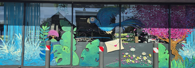 Vue large de la vitrine d'un supermarché décorée d'une fresque colorée représentant un paysage imaginaire avec cascades, oiseau bleu portant une lettre, et végétation luxuriante.